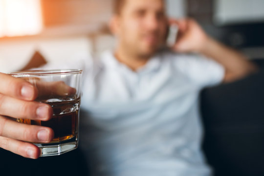 Young man watch tv in his own apartment. Picture with blurred background. Guy talking on phone and hold glass of whiskey in hand. Busy businessman working remotely.
