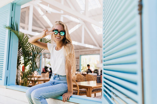 Amazing european female model in white t-shirt posing with peace sign. Portrait of refined blonde girl in sparkle sunglasses and blue jeans.