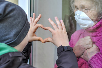 Grandmother mature woman in a respiratory mask communicates with her grandchild through a window....