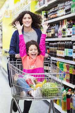 Mother pushing excited daughter in shopping cart in supermarket aisle