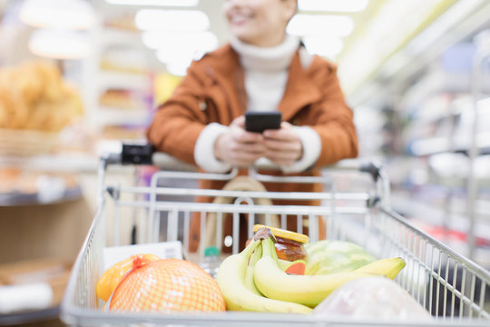 Woman with smart phone pushing shopping cart in supermarket