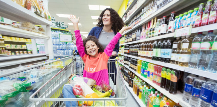 Mother pushing playful daughter in shopping cart in supermarket aisle