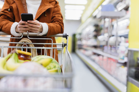 Woman With Smart Phone Pushing Shopping Cart In Supermarket