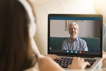 Back view of young girl relax on couch speak on video call on laptop with elderly father, millennial female communicate online use Webcam conference application talk with senior dad from home