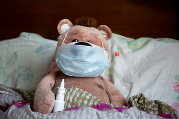a teddy bear lies in bed in a medical mask with a thermometer.