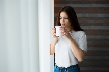 Young woman on kitchen during quarantine. Stand at window and drink coffee or tea. Look outside through curtain. Calm peaceful woman alone in room.