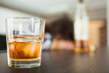 Young woman in kitchen during quarantin. Glass with whiskey or another alcohol drink in front. Sleppy drunk woman with bottle on blurred background.