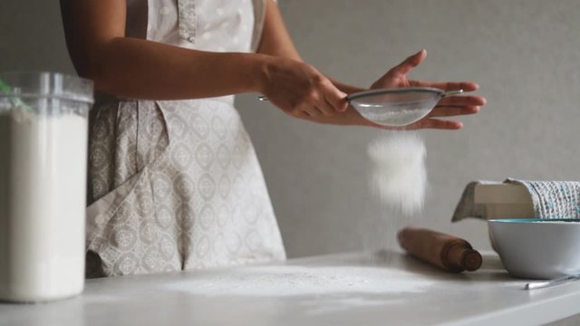 A woman in a beige apron is cooking on the surface of a bright kitchen table. Natural light.  The girl sifts the flour through a sieve on the working surface of the table to work with the dough.