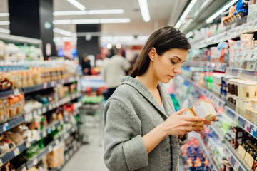 Foto op Plexiglas Young woman shopping in the supermarket grocery store.Reading ingredients,declaration or expiration date on a dairy product before buying it.Nutritional values of the food.Lactose intolerance © eldarnurkovic