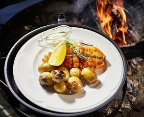 Grilled fish is on white plate with potatoes, lemon and sauce, naked flame, red coals, a smoke, firewood, 