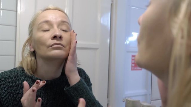 A woman in the bathroom applies a liquid foundation to her face in front of a mirror.
