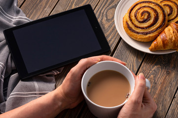 A woman having breakfast on a rustic table checking the latest on her electronic device while having tea and pastry.