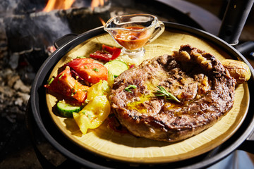 Fototapeta na wymiar The big piece of the grilled meat and vegetables lies on a plate, Juicy beef steak, near big naked flame, sauce, red coals, a smoke, firewood