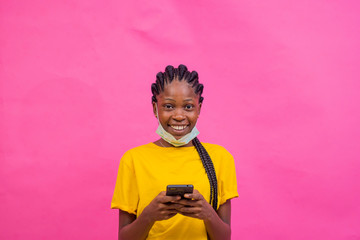 a pretty young female holding her mobile phone while she looks at the camera. african girl wearing a medical face mask.