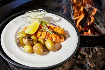 Grilled fish is on white plate with potatoes, lemon and sauce, naked flame, red coals, a smoke, firewood, 