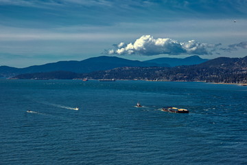 Vancouver harbor, tug-boat tows a barge, ocean tankers are waiting for loading in the port on a sunny windy day against the backdrop of a mountain ridge  and cloudy sky