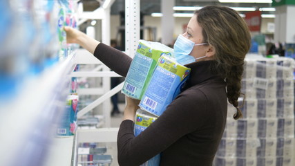 A young woman in a medical mask takes a lot of toilet paper in a supermarket