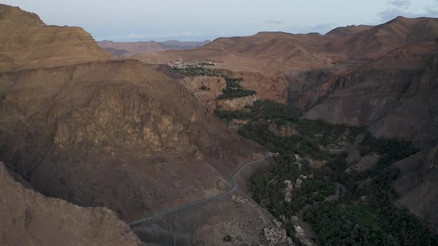 A dusk or dawn aerial push over a lush valley of palms towards a village overlooking a gorge, encircled by a steep headwall of jagged, desert, mountain peaks.