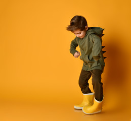 Little brunet kid in khaki dino hoodie with hood and pants, yellow rubber boots. He is posing...