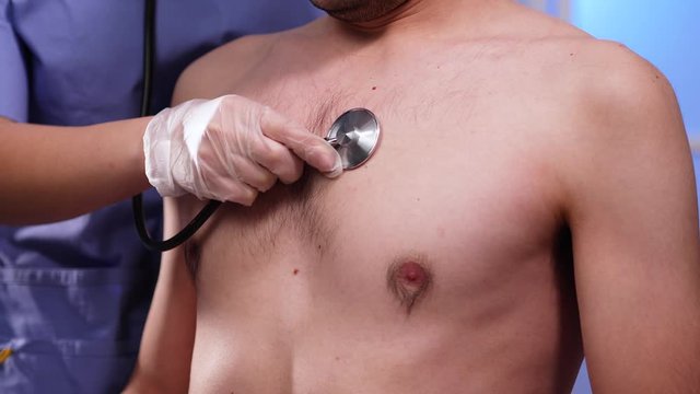 A female woman medical professional doctor/ nurse, wearing PPE protective gloves, scrubs , using a stethoscope to listen to a shirtless male patients lung sounds,on his chest asking him to breath in a