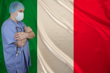 Fototapeta na wymiar male doctor in a medical mask with a stethoscope on the background of the silk national flag of italy, concept of medical care, quarantine, tourism