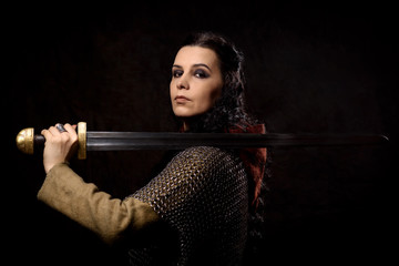 A dark-haired woman with a sword, a Scandinavian warrior in a chain mail against a dark background