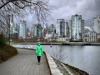 Fototapeta na wymiar Middle age woman in face protective mask walking on seawall during CoVid 19 pandemic alone. View of downtown and Falls Creek. Vancouver. British Columbia. Canada 
