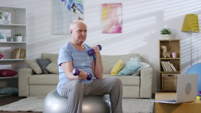 Senior man sitting on exercise ball, watching video workout on laptop and doing dumbbell curls while working out in the living room at home
