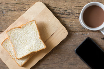 Top view of Breakfast bread and coffee in a white cup and Smart phone on a wooden table with morning sunlight and  copy space