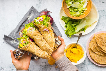 Vegan healthy Mexican tortilla flatbread wraps stuffed with green salad leaves, onion and chickpea hummus filling. Woman hold vegetarian food appetizer in hands. Homemade