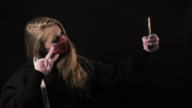 Girl With A White Ribbon And The Inscription Covid 19 In Red Mask Takes Selfie On Smartphone. Isolated On Black Background. Health Care And Medical Concept. Coronavirus Epidemic