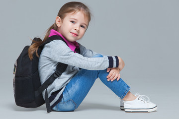Primary school girl in jeans and uniform jacket with backpack is sitting on the floor. Isolated on grey background
