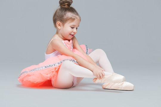 Cute charming ballerina little girl in pink tutu is sitting on the floor trying to put on ballet shoes