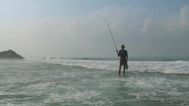 aged Sinhalese fisherman stands in foaming ocean water near beach under cloudy sky in summer. Concept aboriginal people