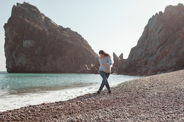 Obraz premium Happy pretty girl walks along the sea coast against the background of the sea and rocks, from behind a beautiful landscape