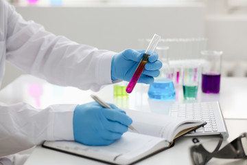 Employee of the chemical laboratory teacher chemist holds a silvery pen in his hand makes notes in the diary records test data from reactions examining test tubes with the substance arm in gloves.