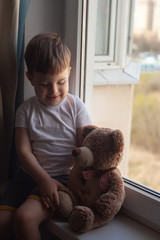 Stay at home quarantine coronavirus prevention of the pandemic. A child and his Teddy bear on the windowsill and look out the window. The view of the street. Prevention of the epidemic.