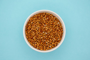 Dry buckwheat in white ceramic bowl isolated on blue. Spilled buckwheat.