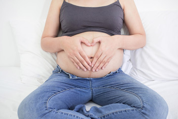 Happy pregnant asian woman sitting on bed and touching her big belly, Heart shape made from hands