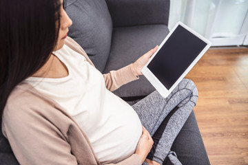 beautiful asian pregnant woman smiling using smart tablet device watching video placing hands on baby lump, sitting on sofa relaxing resting in living room with brick texture wall and white curtains