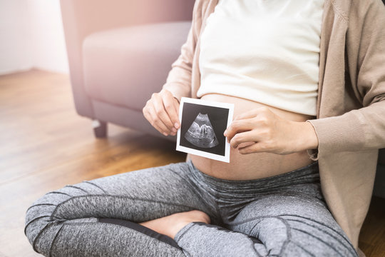 close up asian pregnant woman sitting on floor against sofa holding ultrasound baby picture smiling with happiness resting relaxing in living room from stress wearing comfy stretch pants and cardigan