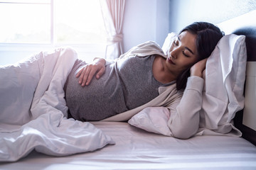 Beautiful asian pregnant woman laying down on bed sideways sleeping taking a nap with pregnant belly out, resting and relaxing in bed room from hormone stress, comfy wearing stretch pants and cardigan