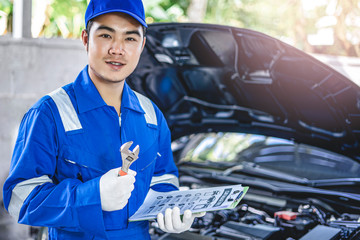 Concept of male asian car mechanic smiling holding a wrench and a checkup list of car engine, checking up on the car engine, for repair and checkup, wearing white gloves and a blue overall for safety