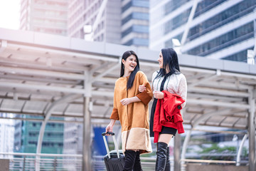 beautiful two asian women dressed casually holding each other arms smiling and laughing joyfully, holding a jacket in arm and dragging suitcase, walking through the urban city structure in background