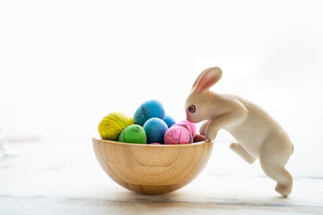 Fototapeta na wymiar isolated vibrant colorful easter egg green, blue, yellow and pink in a wooden bowl with toy white nummy rabbit hopping on the bowl on a wooden table with white background, representing easter holidays