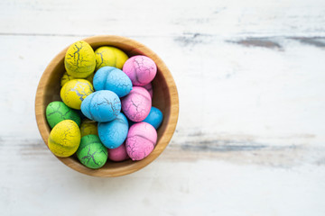 flat lay colorful vibrant easter egg of pink green yellow and blue colors piling in brown wooden bowl, on wooden table with white scraped paint background, representing celebration of easter holidays