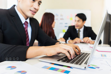 closeup of businessman working on the laptop computer with hands typing on the keyboard, working in an office environment with notebook, documents and colleagues wearing suit and tie in the background