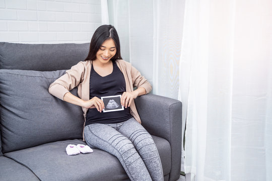 beautiful asian pregnant woman sitting on sofa holding ultrasound baby picture while placing hand on belly resting relaxing in living room from hormone stress, wearing comfy stretch pants and cardigan