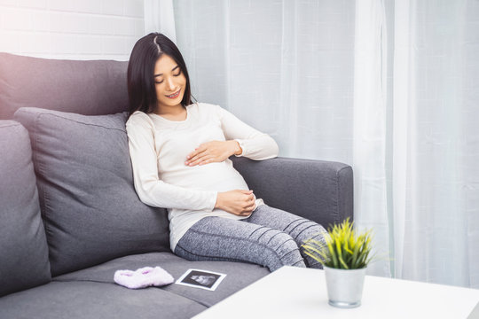 beautiful asian pregnant woman placing hands on baby lump feeling heartbeat smiling joyfully, sitting on sofa relaxing resting from tiredness, in living room with brick texture wall and white curtains