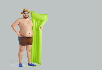Fat funny man in swimming trunks with an inflatable mattress on a gray background.